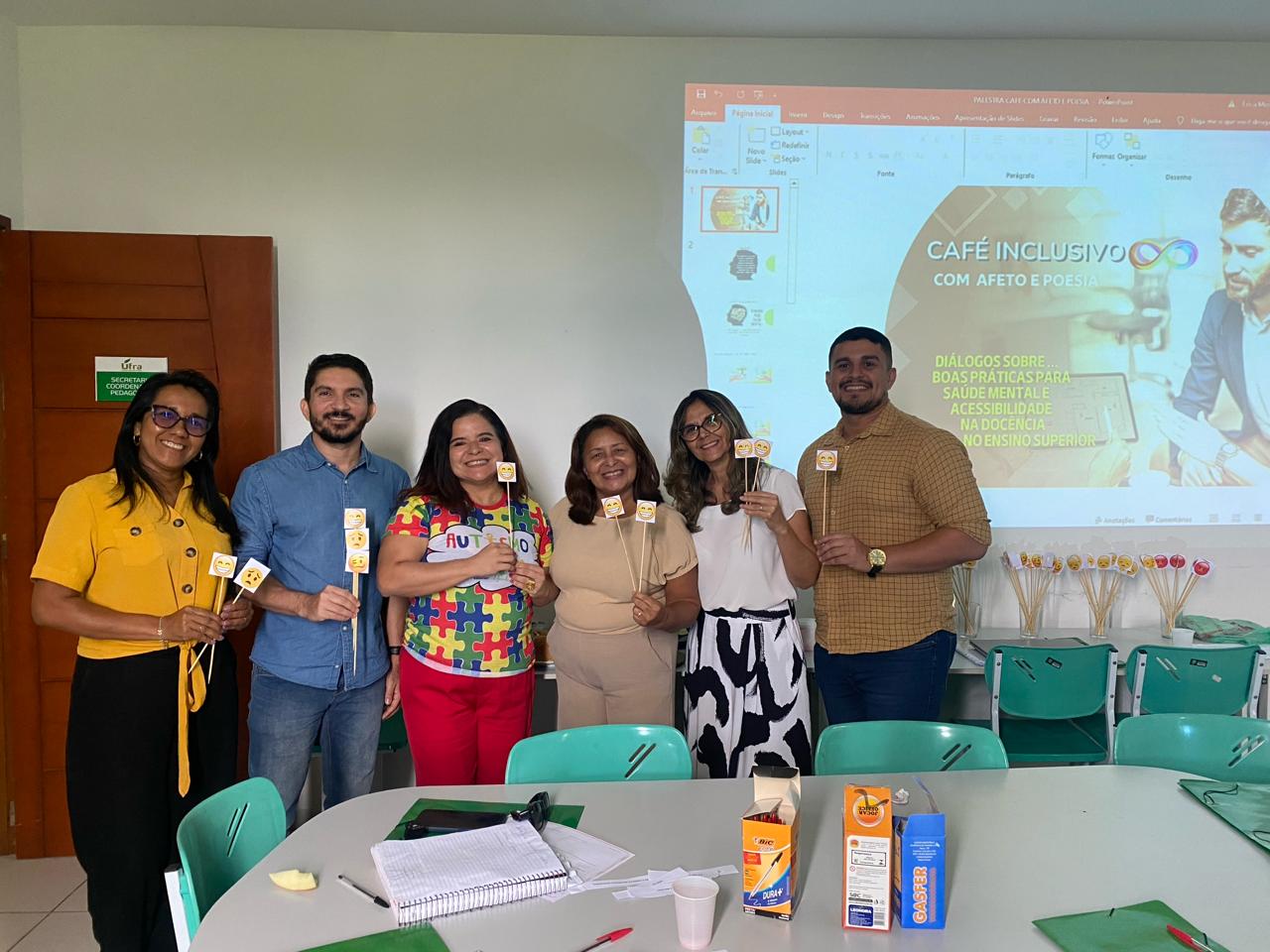 Imagem de cinco pessoas entre homens e mulheres, estão em pé e posam sorridentes segurando imagens de emoji em palito. Estão em uma sala, com mesas e cadeiras à sua frente, atrás reflete material de apresentação em uma parede branca